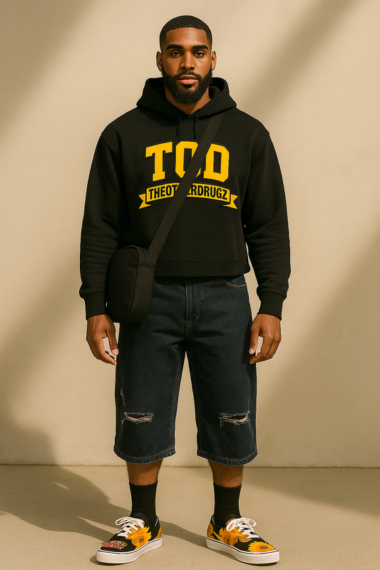 Man wearing a black hoodie with yellow text, dark jeans, and sneakers on a beige background